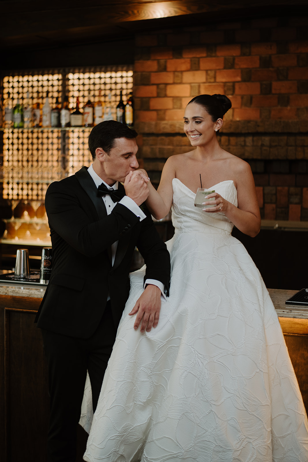 Groom kissing the bride's hand as she sits on the bar counter holding a cocktail.